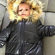 Shelsea participe au concours pour gagner de l'argent avec cette photo : baby, child, puffer_jacket, fur_hood, black_clothing, couch, indoor, infant, warm_clothing, face, wide_eyes, soft_furniture, winter_wear, zipper, small_person, portrait, expression, seated, comfort, curious