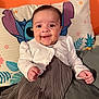Bella is registered to the contest to win money with this photo: baby, smiling, child, cardigan, dress, pleated, pillow, stitch, couch, orange_wall, happy, indoors, portrait, cute, small_hands, clothing, face, seated, infant, cozy