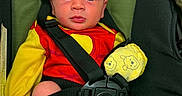 Alban is registered to the contest to win money with this photo: baby, infant, car_seat, seat_belt, clothing, mittens, red_outfit, yellow_sleeves, face, eyes, head, person, child, sitting, safety, straps, cute, indoors, portrait, expression