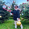 child, toddler, outdoor, christmas_tree, ornament, holiday, winter_clothing, grass, festive, toy, smiling, coat, hood, mittens, pants, shoes, nature, daylight, celebration, cute