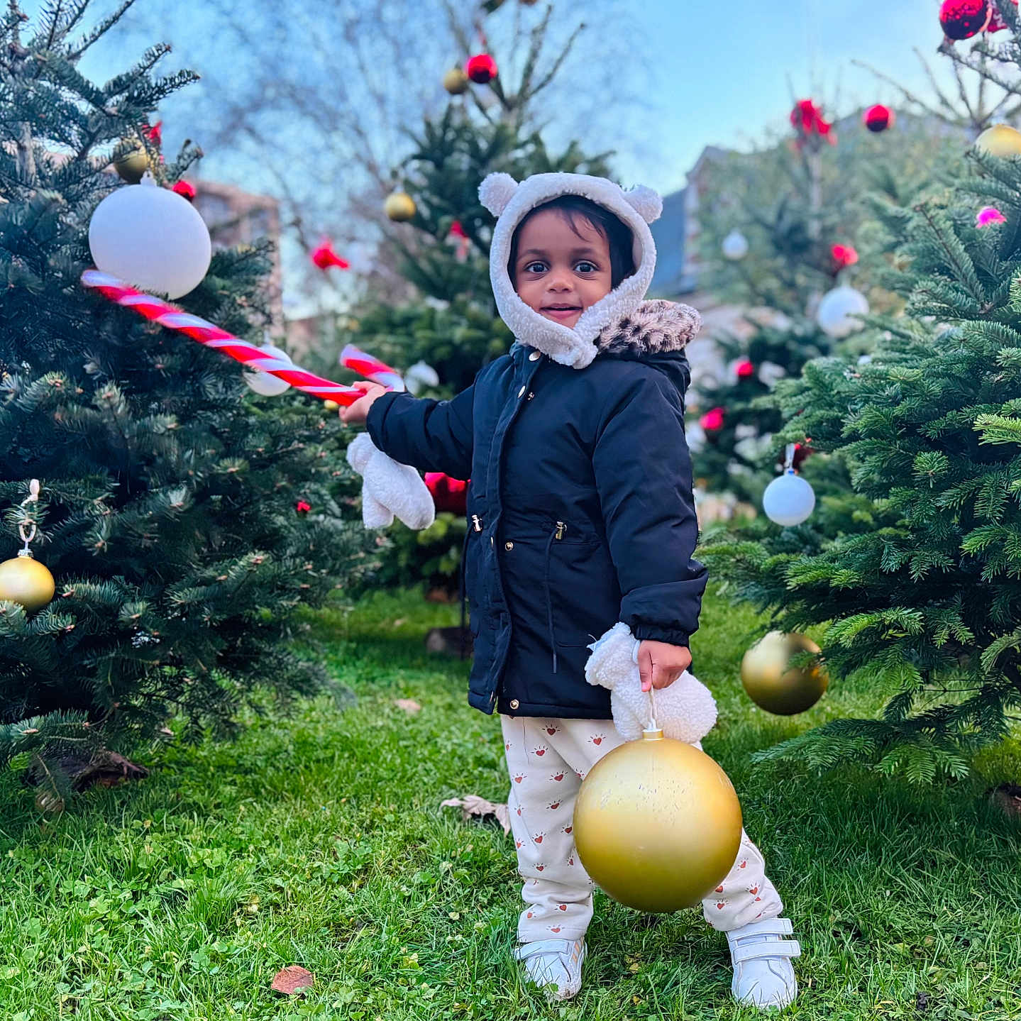 Jaya a rejoint le concours — aidez-le/la à gagner de superbes lots ! child, toddler, outdoor, christmas_tree, ornament, holiday, winter_clothing, grass, festive, toy, smiling, coat, hood, mittens, pants, shoes, nature, daylight, celebration, cute