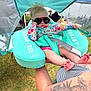 Elena is registered to the contest to win money with this photo: baby, sunglasses, floatation_device, tattoo, arm, outdoor, grass, colorful_clothing, infant, child, person, summer, festival, shade, sitting, relaxing, sun_protection, baby_seat, floral_pattern, casual