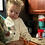 baby, child, headband, shirt, person, indoor, kitchen, cabinet, coffee_maker, countertop, water_bottle, hand, holding, facial_expression, clothing, appliance, container, wood, face, young