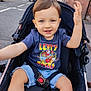 toddler, child, stroller, street, urban, casual_clothing, shorts, tshirt, smiling, waving, daylight, sidewalk, car, safety_belt, playful, outdoor, person, young_child, cute, happy