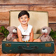 Ezekyel participe au concours pour gagner de l'argent avec cette photo : toddler, child, smiling, suitcase, plush_toys, teddy_bears, wooden_floor, wooden_background, green_leaves, vintage, indoor, portrait, happy, cute, sitting, clothing, white_shirt, yellow_shorts, playful, fun
