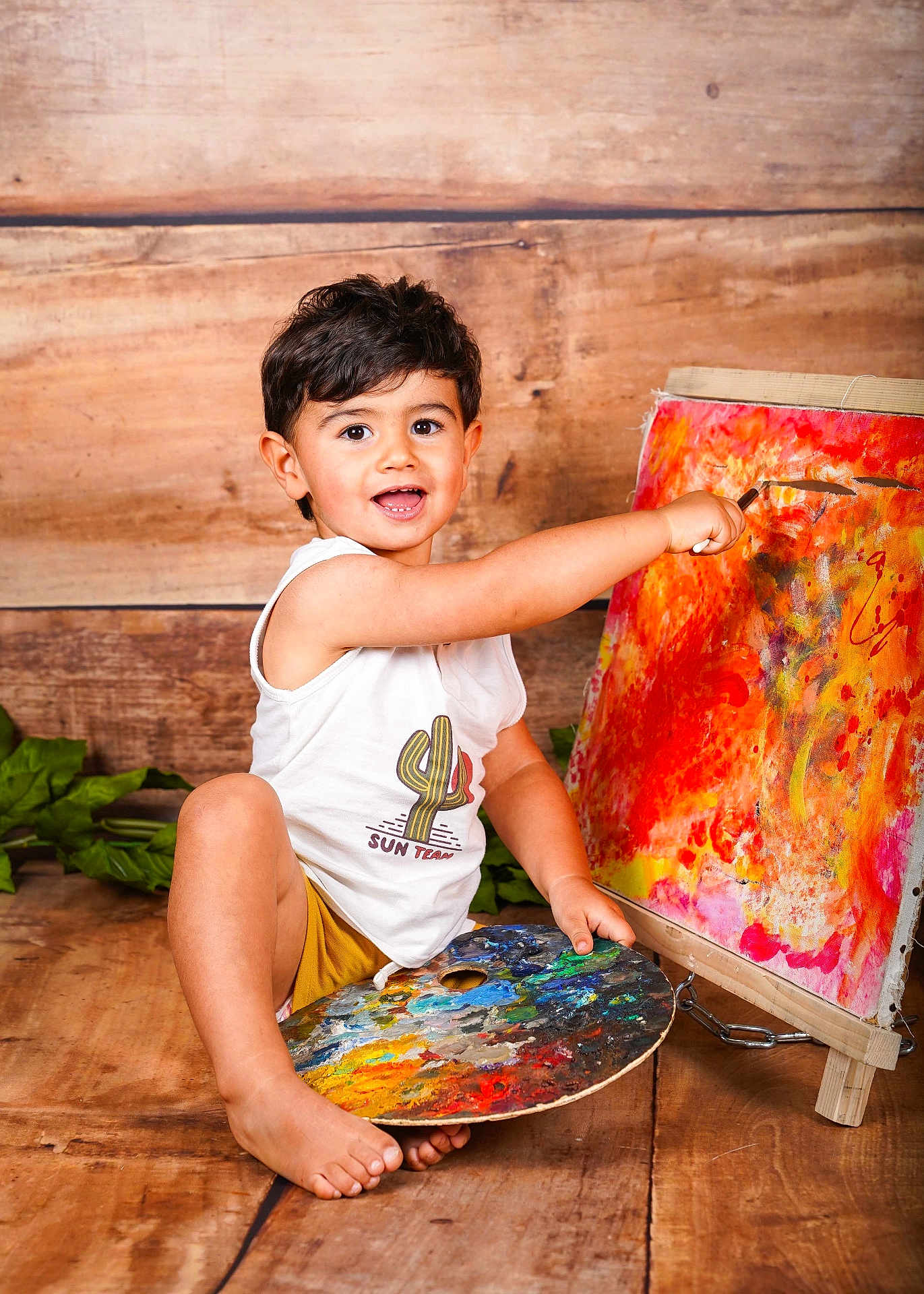Ezekyel a rejoint le concours — aidez-le/la à gagner de superbes lots ! child, toddler, painting, art, palette, canvas, smiling, happy, barefoot, wooden_floor, white_tank_top, yellow_shorts, indoors, creative, colorful, expression, sitting, person, brush, playful