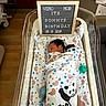 Sonny is registered to the contest to win money with this photo: newborn, baby, hospital, bassinet, blanket, panda, sleeping, letter_board, text, swaddled, infant, pacifier, medical_equipment, wood_floor, couch, pillows, wrapped, cute, child, bed