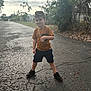 child, boy, standing, outdoor, road, asphalt, cracked_surface, orange_shirt, black_shorts, sneakers, gold_chain, smiling, confident, daylight, nature, trees, plants, casual_clothing, young, person