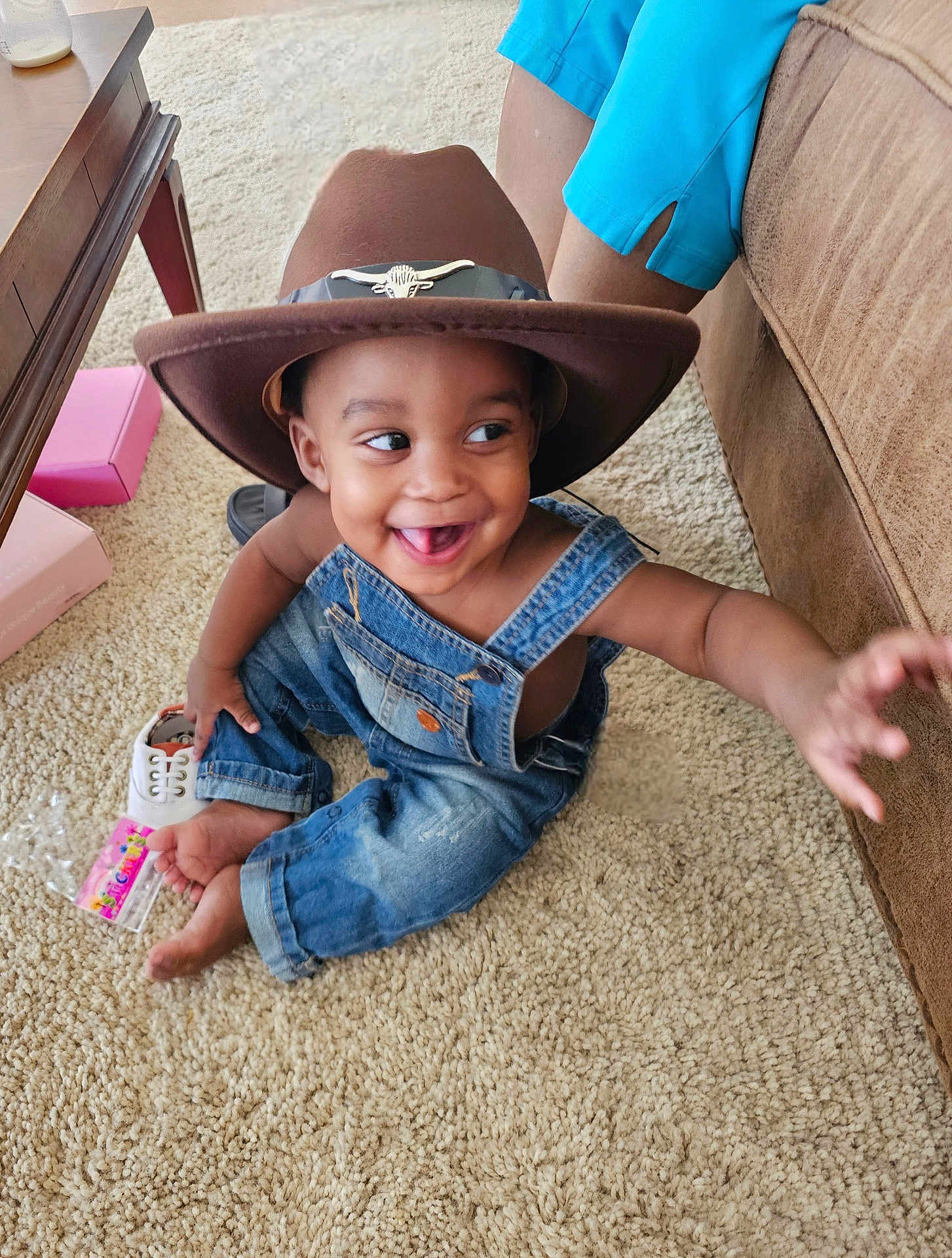 Justin joined the competition — help win amazing prizes! toddler, child, hat, cowboy_hat, denim_overalls, carpet, smiling, happy, indoor, person, footwear, toy, hand, couch, legs, blue_shorts, wooden_table, box, barefoot, playful