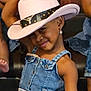 child, denim_dress, cowboy_hat, smile, earrings, pose, playful, fashion, girl, cute, portrait, indoor, wooden_background, accessory, happy, young, person, denim, jewelry, fun
