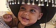 Layla is registered to the contest to win money with this photo: baby, smiling, hat, sombrero, beads, child, cute, indoors, blanket, pattern, face, portrait, happy, infant, clothing, person, young_child, skin, earrings, black_top