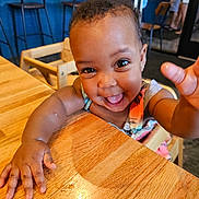 Layla is registered to the contest to win money with this photo: toddler, child, smiling, happy, reaching, table, wooden_table, indoor, high_chair, person, face, eyes, arm, clothing, restaurant, background, people, furniture, baby, cute
