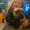 child, dog, french_bulldog, hugging, indoor, couch, blue_wall, furniture, portrait, smiling, curly_hair, cozy, home, person, pet, casual_clothing, table, room, yellow_cabinet, photography