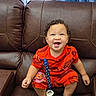 baby, child, toddler, smiling, happy, curly_hair, portrait, sitting, leather_couch, recliner, orange_outfit, racing_theme, number_95, sandals, pacifier, blue_curtains, indoor, home, fashion, photography
