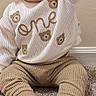 baby, child, toddler, smiling, tongue_out, curly_hair, portrait, sitting, indoor, carpet, sweater, bear_pattern, knitwear, pants, shoes, brown_shoes, raised_arm, home, wall, cute