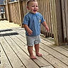 toddler, child, barefoot, blue_tshirt, gray_shorts, wooden_deck, railing, outdoor, smiling, happy, standing, wood, person, casual_clothing, daylight, young_child, expression, portrait, natural_light, fun