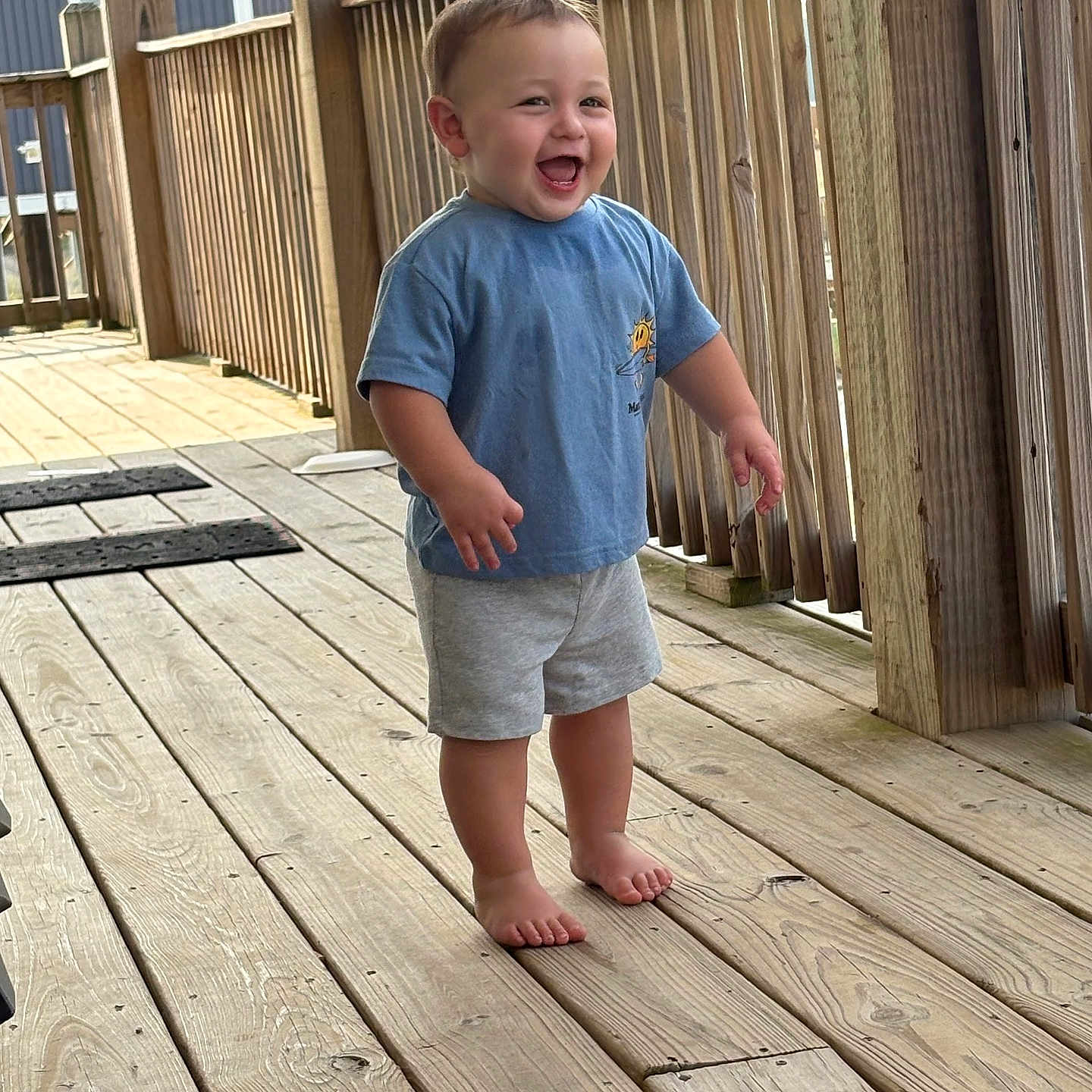 Aiden joined the competition — help win amazing prizes! toddler, child, barefoot, blue_tshirt, gray_shorts, wooden_deck, railing, outdoor, smiling, happy, standing, wood, person, casual_clothing, daylight, young_child, expression, portrait, natural_light, fun