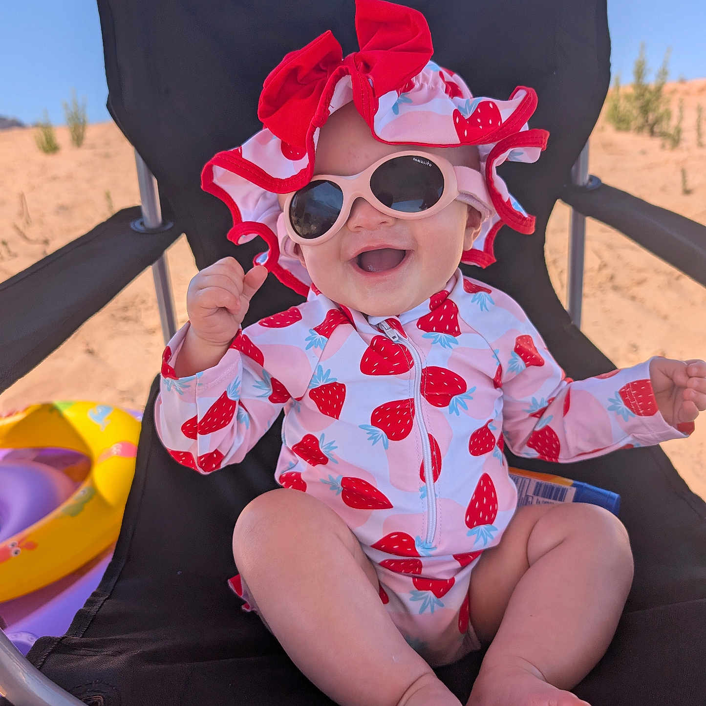 Jade is registered to the contest to win money with this photo: baby, beach, beach_chair, child, clothing, feet, happy, hat, inflatable_toy, outdoor, relaxation, sand, seat, smile, strawberry_pattern, summer, sunglasses, sunny, swimwear, toddler
