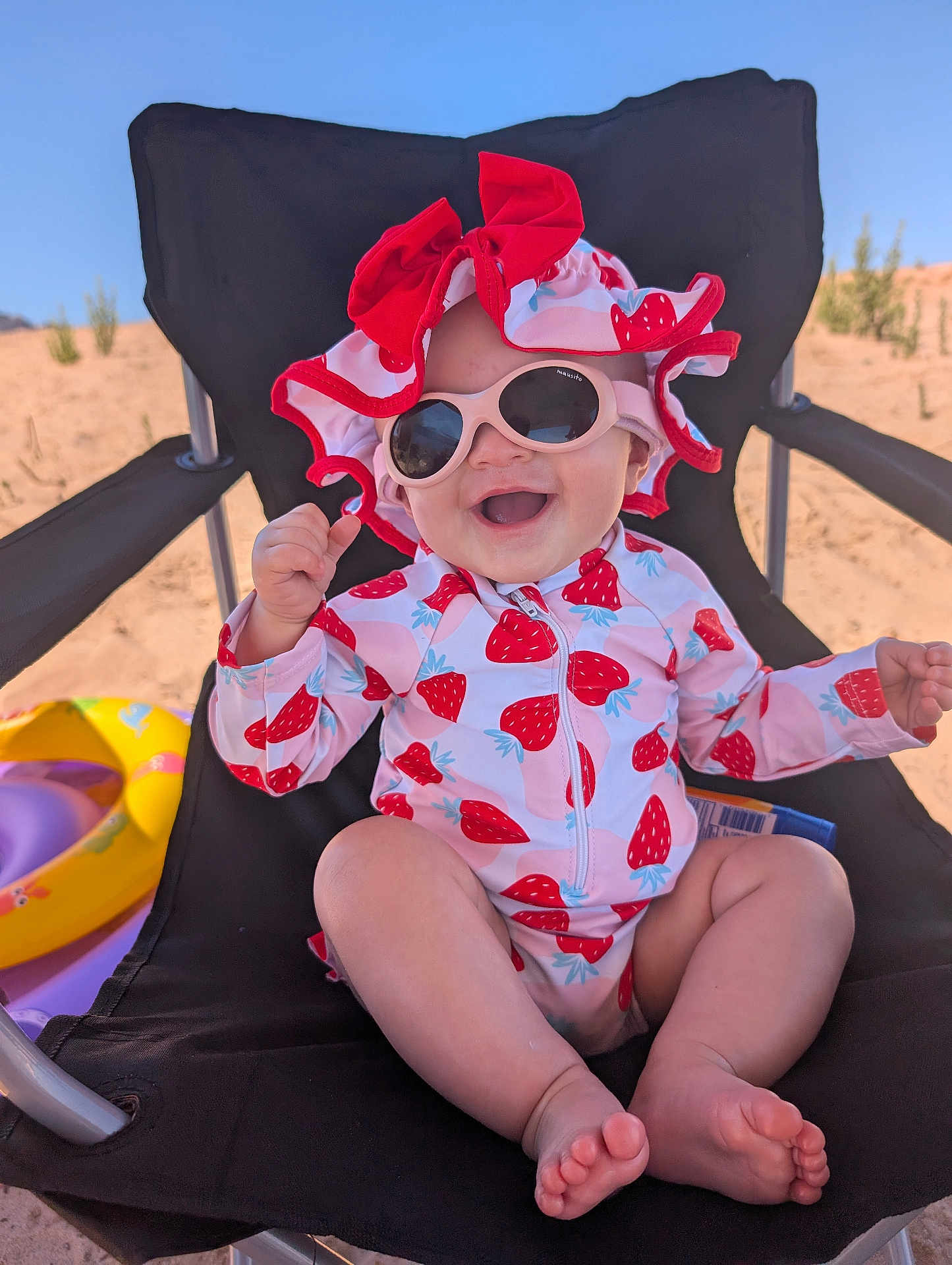 Jade is registered to the contest to win money with this photo: baby, child, sunglasses, hat, strawberry_pattern, swimwear, beach_chair, beach, sand, inflatable_toy, smile, happy, sunny, outdoor, summer, toddler, feet, clothing, seat, relaxation