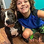 child, dog, smiling, curly_hair, blue_shirt, green_shorts, barefoot, wooden_floor, indoor, pet, happy, portrait, black_dog, white_markings, teal_wall, crouching, friendship, cute, animal, home