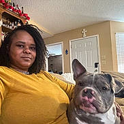 Nipsey joined the competition — help win amazing prizes! bottles, brown_skin, casual, companion, couch, curly_hair, dog, door, furniture, home, indoor, living_room, pet, portrait, relaxing, seated, shelf, window_blinds, woman, yellow_sweater