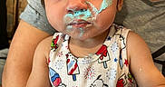 Kali is registered to the contest to win money with this photo: toddler, child, cream, face_paint, blue_tutu, colorful_clothing, indoor, person, sitting, cute, expression, closeup, smile, funny_face, baby, arm, clothing, cartoon_print, food_mess, parent