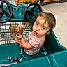 toddler, child, shopping_cart, cookie, grocery_store, toy_car, steering_wheel, floor, wooden_shelves, clothing, brown_hair, indoor, seat, looking_up, curious, small_child, sitting, hand_holding, expression, retail