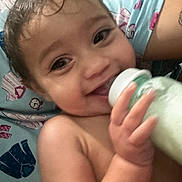 Kali is registered to the contest to win money with this photo: baby, smiling, milk_bottle, hand, face, wet_hair, child, bed, blanket, patterned_bed_sheet, cute, infant, skin, person, happy, portrait, indoors, young_child, feeding, closeup