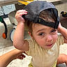 toddler, child, hat, cap, mesh_hat, indoors, floor, hand, yellow_clothing, cream_top, curious_expression, sitting, baby, person, headwear, young_child, casual, home, portrait, cute