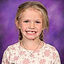 child, girl, smiling, portrait, braids, missing_tooth, necklace, floral_pattern, long_sleeves, blue_eyes, blonde_hair, studio_background, purple_background, happy, cute, school_photo, casual_clothing, young, female, face