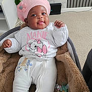 Ayana joined the competition — help win amazing prizes! baby, infant, pink_hat, flower, white_clothing, cartoon_cat, blanket, baby_carrier, smiling, indoor, cozy, child, cute, plush, toy, pacifier, carpet, furniture, person, portrait