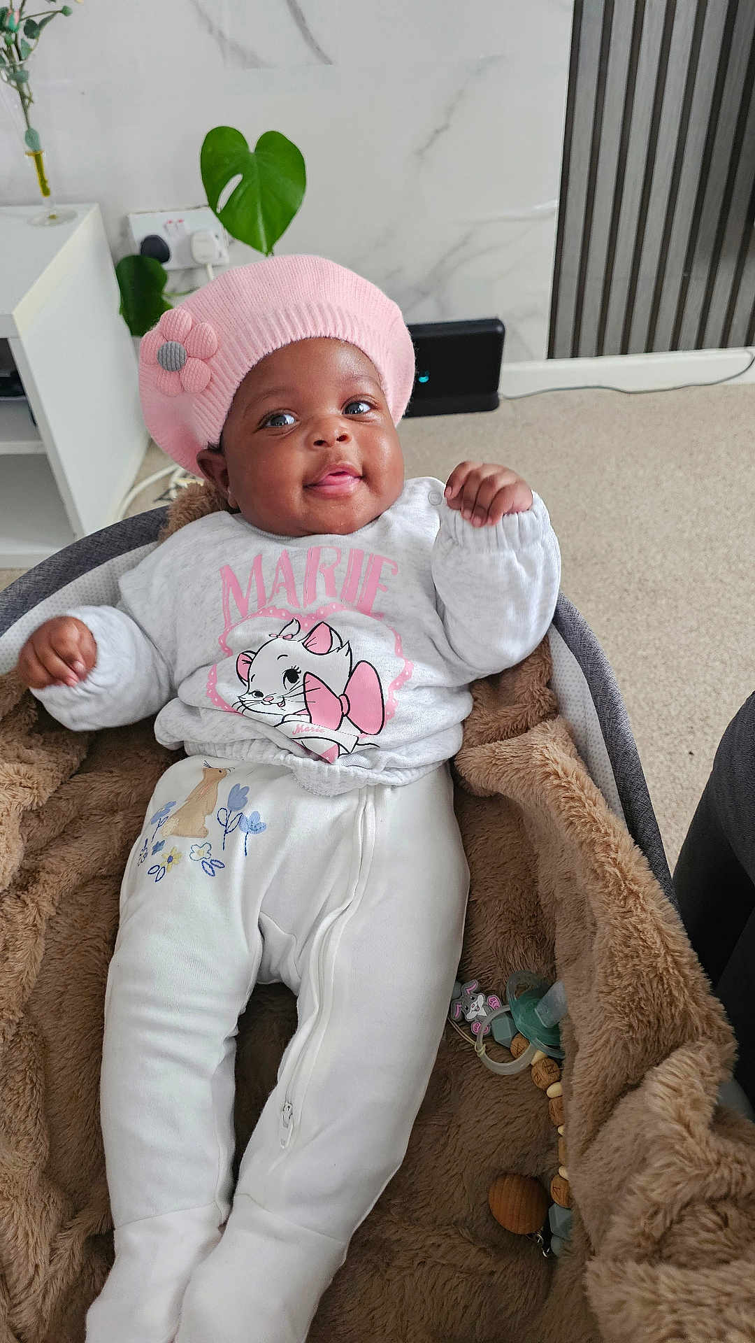 Ayana joined the competition — help win amazing prizes! baby, infant, pink_hat, flower, white_clothing, cartoon_cat, blanket, baby_carrier, smiling, indoor, cozy, child, cute, plush, toy, pacifier, carpet, furniture, person, portrait