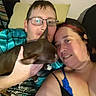 bed, black_clothing, blanket, brown_dog, couch, couple, cuddling, dog, glasses, home, indoors, man, person, pet, pillow, portrait, relaxed, selfie, smiling, woman