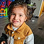 child, smiling, indoor, playroom, toy, television, hazel_eyes, jacket, brown_hair, buttoned_clothing, floor, wooden_floor, furniture, colorful, happy, portrait, young_child, person, casual_clothing, playtime
