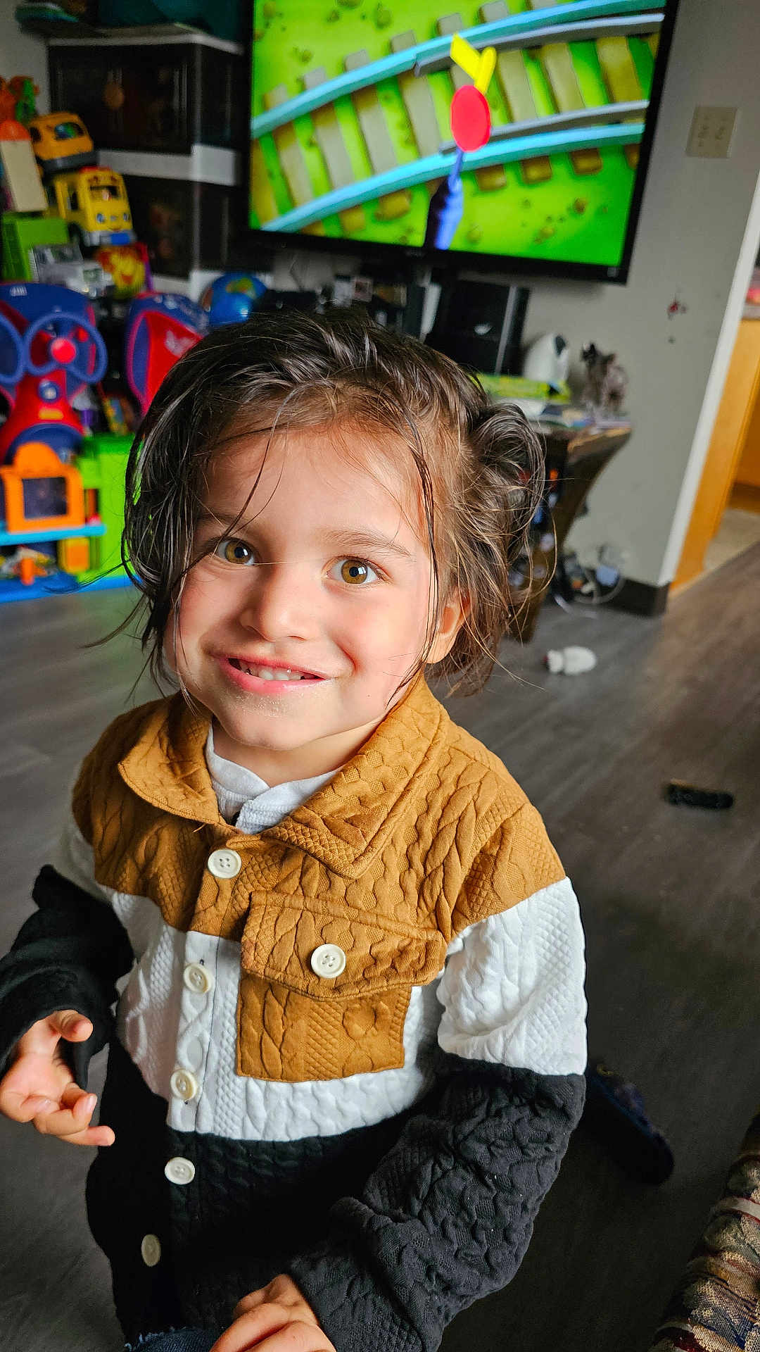 Brentley is registered to the contest to win money with this photo: child, smiling, indoor, playroom, toy, television, hazel_eyes, jacket, brown_hair, buttoned_clothing, floor, wooden_floor, furniture, colorful, happy, portrait, young_child, person, casual_clothing, playtime