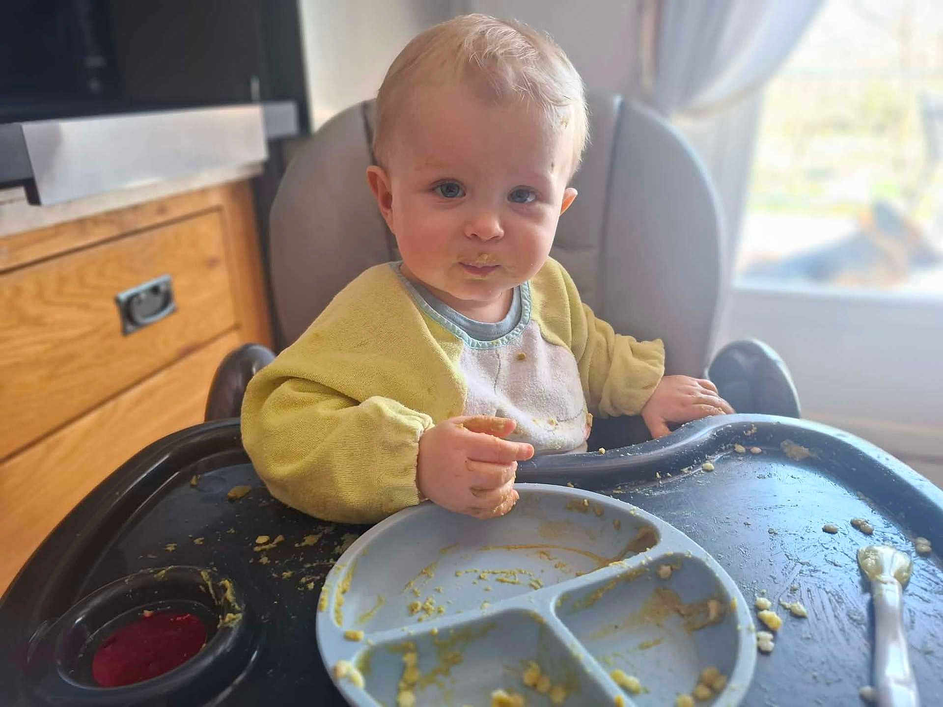 Enzo a rejoint le concours — aidez-le/la à gagner de superbes lots ! baby, toddler, highchair, bib, messy, baby_food, plate, spoon, eating, mealtime, kitchen, window, sunlight, portrait, blonde_hair, blue_eyes, tray, food_smear, wooden_cabinet, indoor