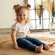 Lylia a rejoint le concours — aidez-le/la à gagner de superbes lots ! child, toddler, barefoot, jeans, white_tshirt, smiling, sitting, indoor, rug, hair_buns, natural_light, window, decor, vase, pampas_grass, wooden_sign, floor, cute, portrait, home