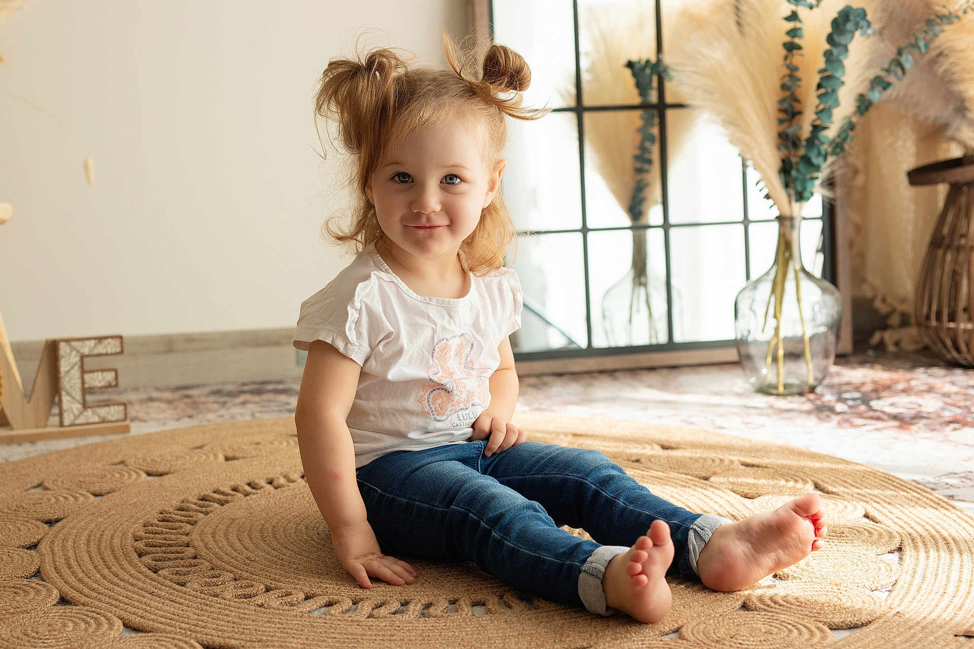 Lylia a rejoint le concours — aidez-le/la à gagner de superbes lots ! child, toddler, barefoot, jeans, white_tshirt, smiling, sitting, indoor, rug, hair_buns, natural_light, window, decor, vase, pampas_grass, wooden_sign, floor, cute, portrait, home