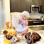 Liam is registered to the contest to win money with this photo: baby, child, chef_hat, rolling_pin, pastries, donuts, cake, kitchen, countertop, flour, baking, smiling, celebration, number_seven, appliance, oven, microwave, bread, happy, person