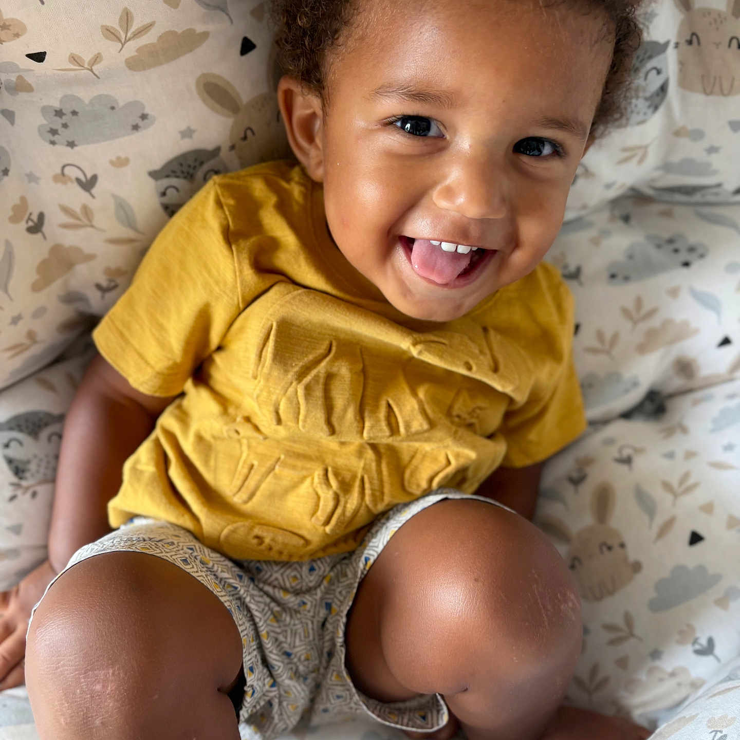 Habib a rejoint le concours — aidez-le/la à gagner de superbes lots ! animal_print, baby, child, cloud_print, curly_hair, cushion, cute, face, happy, indoor, legs, patterned_clothing, playful, portrait, seated, shorts, skin, smiling, toddler, yellow_shirt