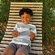 Hamet participe au concours pour gagner de l'argent avec cette photo : child, boy, smile, curly_hair, bench, outdoor, nature, greenery, leaves, wooden_bench, casual_clothing, shorts, tshirt, relaxed, happy, summer, daylight, person, portrait, playful