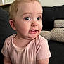 baby, child, infant, person, face, pink_shirt, ribbed_clothing, couch, cushion, indoor, curious_expression, blue_eyes, short_hair, seated, soft_lighting, closeup, skin, mouth, ear, cheek