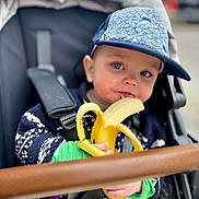 George joined the competition — help win amazing prizes! toddler, child, banana, stroller, cap, blue_cap, sweater, food, snack, cute, baby, outdoor, seatbelt, curious, face, hand, person, closeup, portrait, cozy