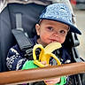 toddler, child, banana, stroller, cap, blue_cap, sweater, food, snack, cute, baby, outdoor, seatbelt, curious, face, hand, person, closeup, portrait, cozy
