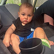 Andrew joined the competition — help win amazing prizes! toddler, child, car_seat, vehicle_interior, black_shirt, blue_shorts, seatbelt, car_door, forest_background, serious_expression, short_hair, skin, hand, footwear, car_window, person, indoors, seat, grip, sitting