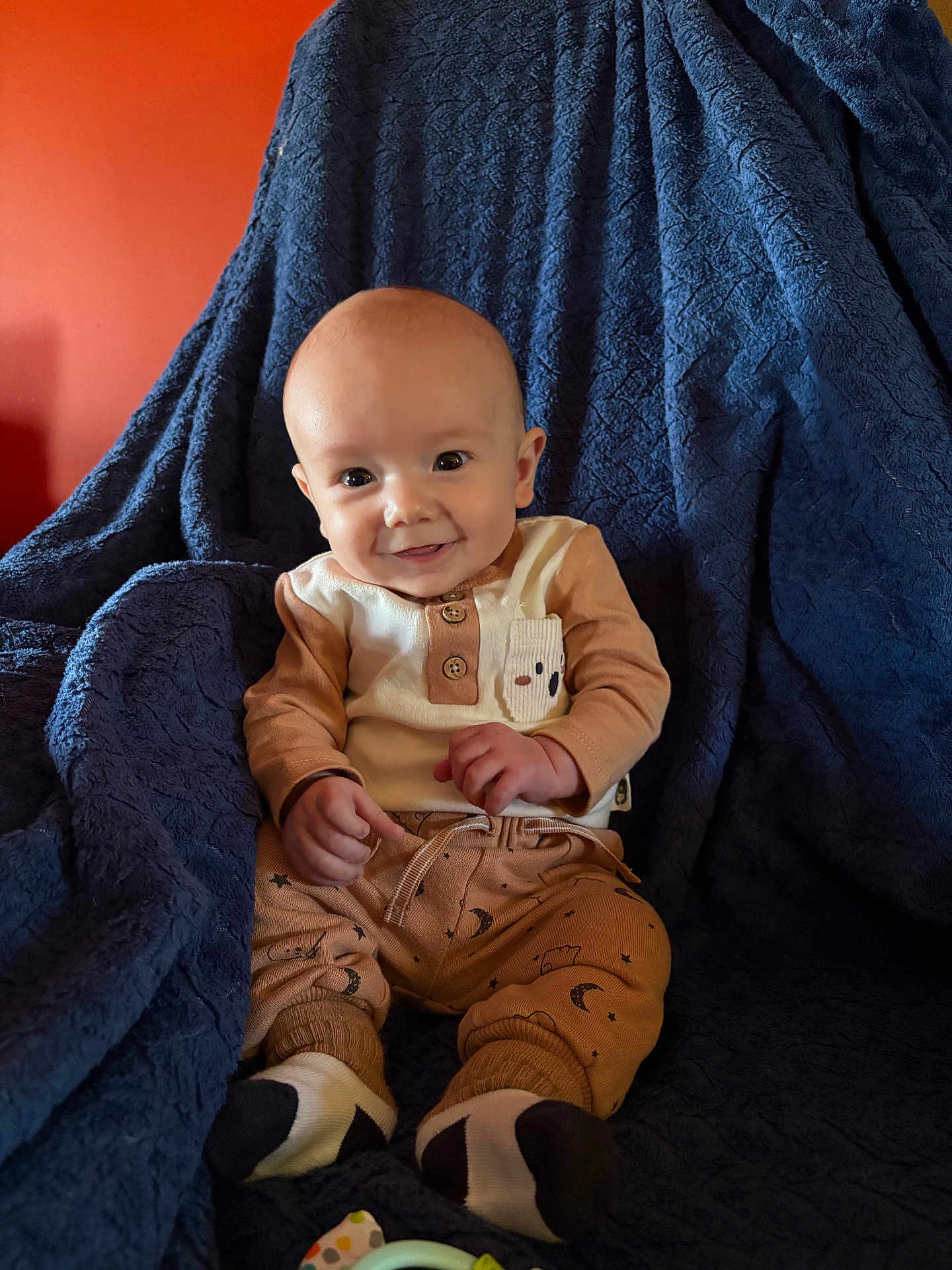 Rowdy is registered to the contest to win money with this photo: baby, child, smiling, sitting, blue_blanket, orange_wall, cozy, cute_clothing, indoor, portrait, happy, infant, face, hands, feet, buttoned_shirt, patterned_pants, warm_lighting, soft_texture, comfortable