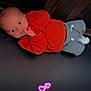 baby, infant, child, orange_sweater, grey_pants, lying_down, black_surface, hand_near_mouth, curious_expression, glowing_hearts, pink_hearts, casual_clothing, cute, small_hands, indoors, portrait, young_child, cozy, resting, love