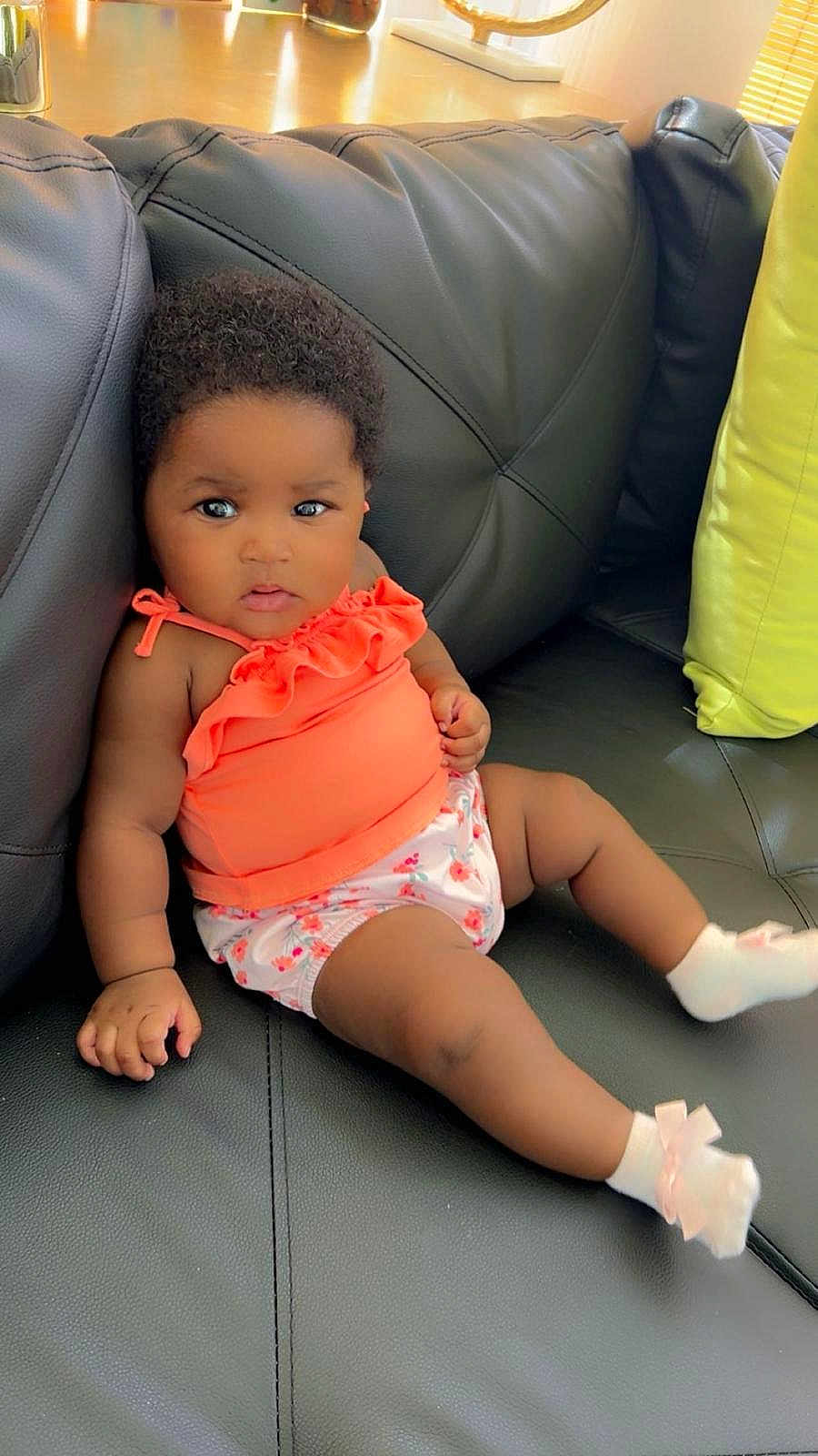 Armony Cara Francis is registered to the contest to win money with this photo: baby, infant, child, couch, black_leather, orange_top, floral_shorts, socks, pink_bows, indoor, furniture, cute, portrait, relaxed, seated, curious, skin, hair, small_hands, expression