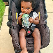 Kevin is registered to the contest to win money with this photo: toddler, child, baby, stroller, sippy_cup, outdoor, sidewalk, grass, curly_hair, seatbelt, drink_holder, foot, hand, clothing, person, infant, sunlight, expression, cute, portrait