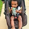 toddler, child, baby, stroller, sippy_cup, outdoor, sidewalk, grass, curly_hair, seatbelt, drink_holder, foot, hand, clothing, person, infant, sunlight, expression, cute, portrait